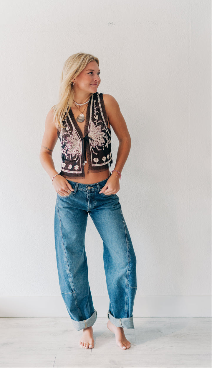A woman stands barefoot against a plain white wall wearing a sleeveless black cropped vest with white floral embroidery and blue wide-leg jeans.