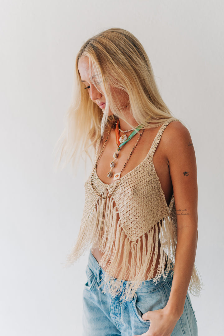 A woman with blonde hair wearing a beige crochet fringed top and light blue denim jeans, standing against a plain white background.