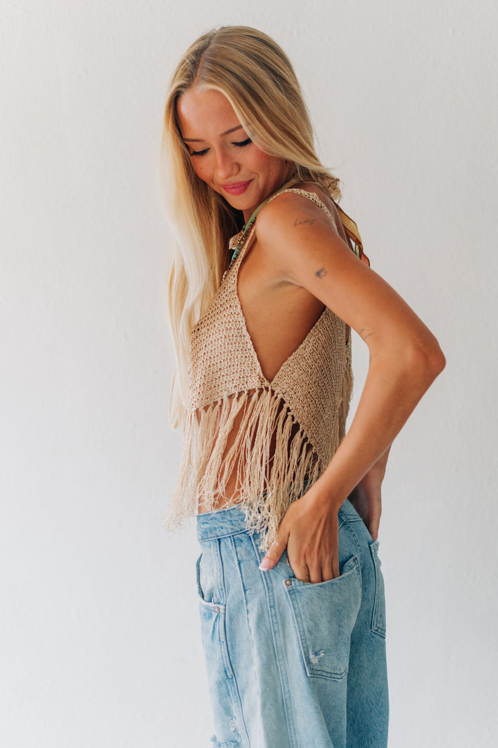 A woman wearing a beige open-knit fringed halter top and light-wash blue jeans, standing against a plain white wall.