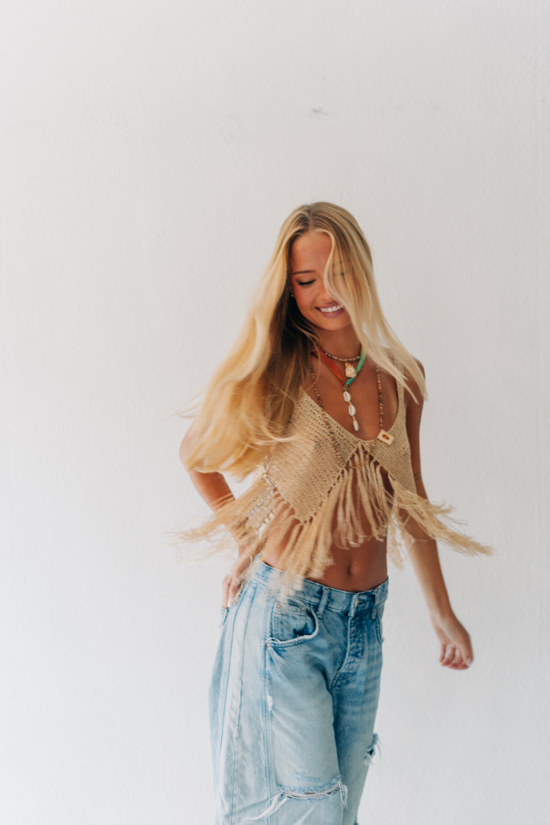 A woman with long blonde hair wearing a beige crochet fringe top and light-wash blue jeans, standing against a plain white wall.