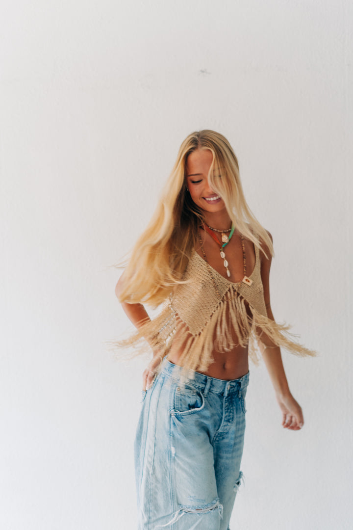 A woman with long blonde hair wearing a beige crochet fringe top and light-wash blue jeans, standing against a plain white wall.