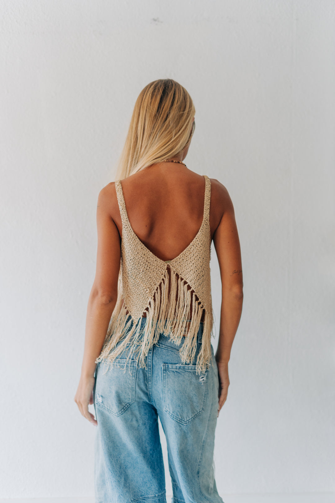 A woman viewed from behind wearing a beige woven fringed backless top and light blue jeans against a plain white wall.