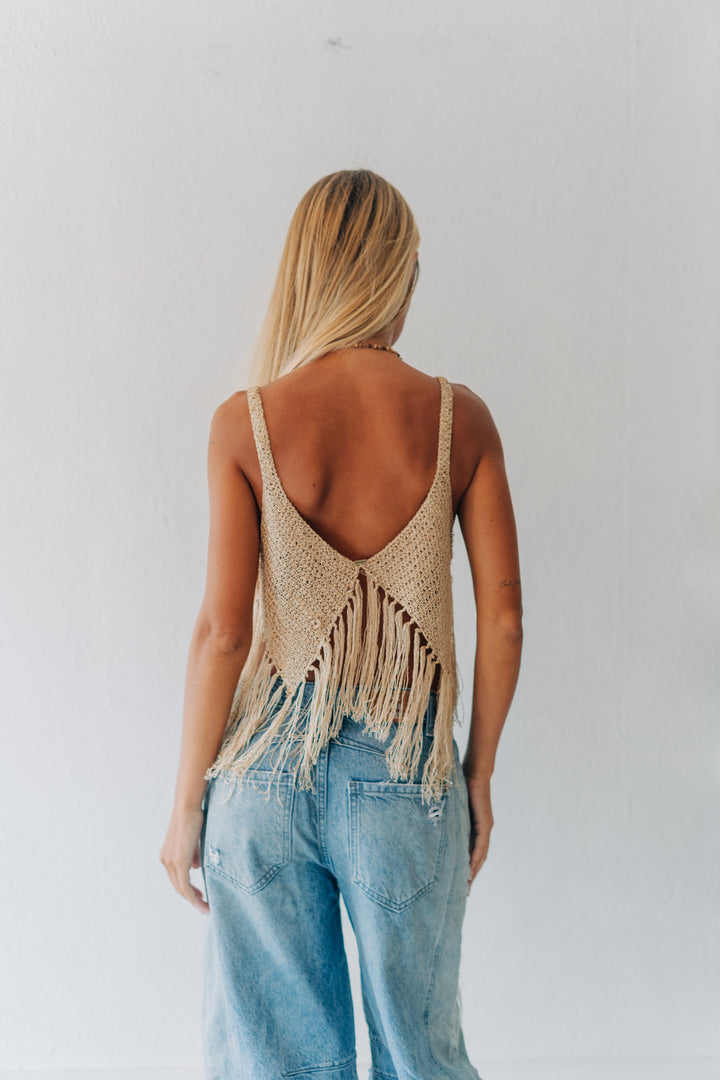 A woman viewed from behind wearing a beige woven fringed backless top and light blue jeans against a plain white wall.