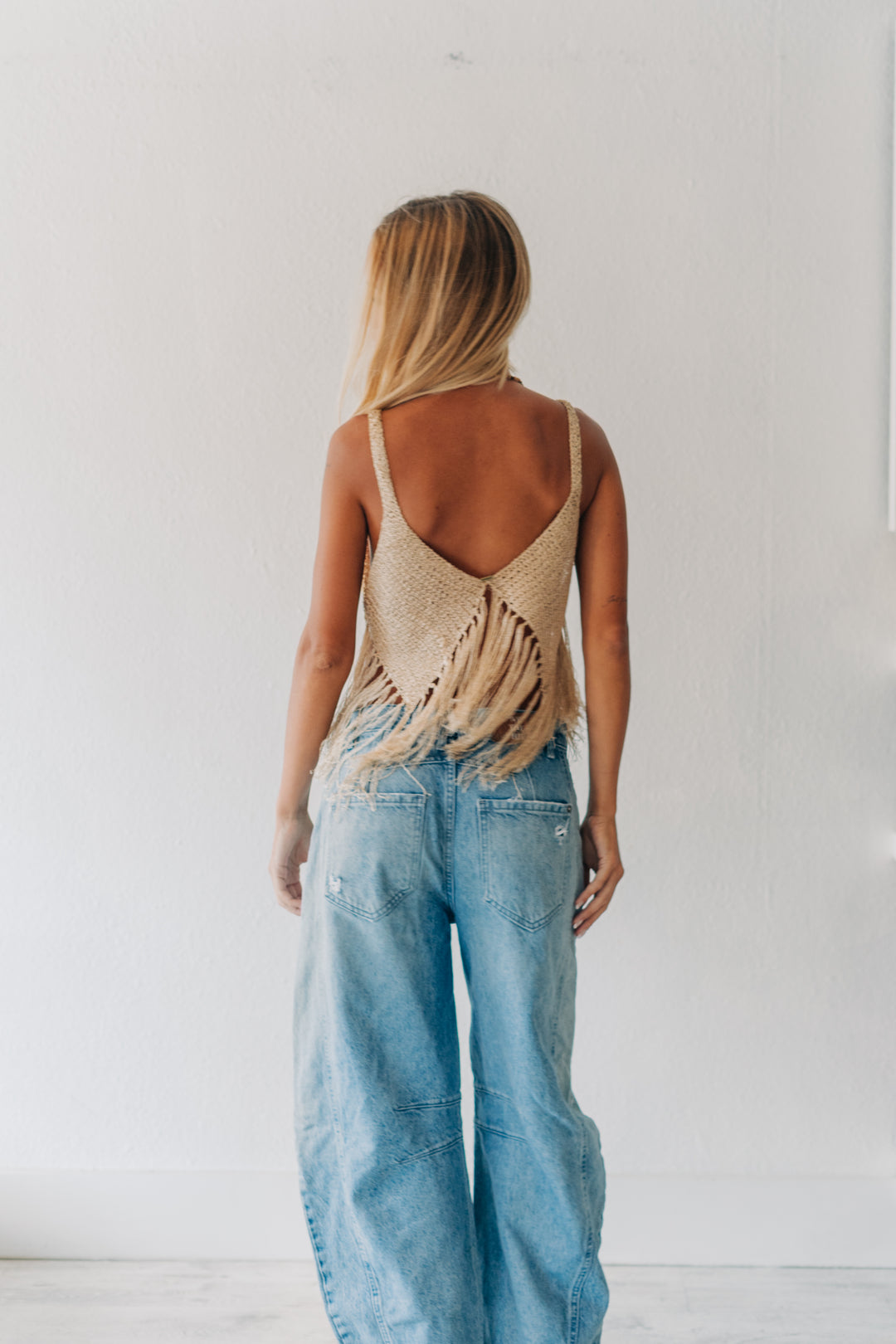 A woman viewed from behind wearing a beige woven open-back top with fringe details and light-wash wide-leg denim jeans, standing against a plain white wall.