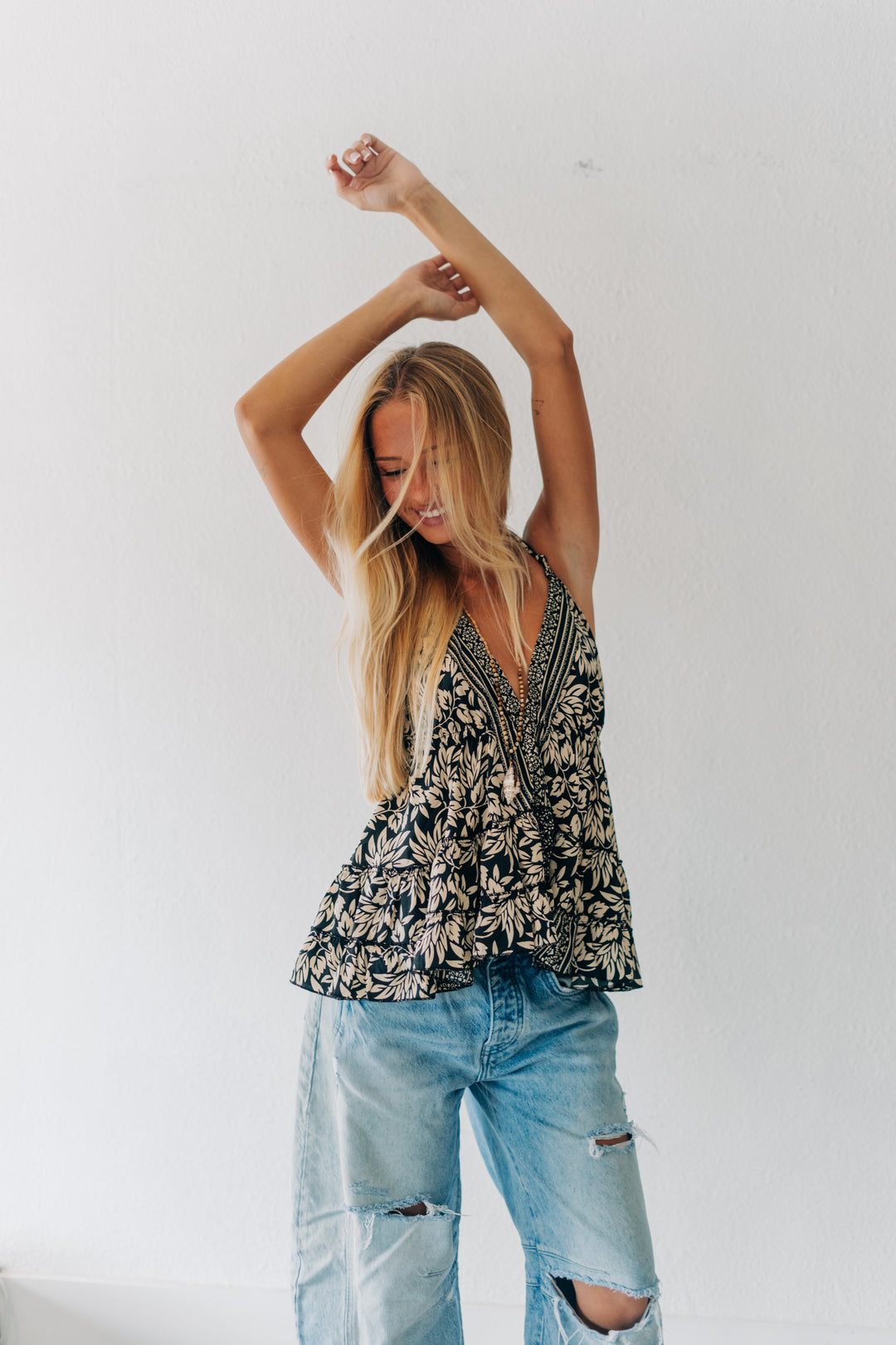 A woman with long blonde hair poses in a black and white botanical print sleeveless top and light-wash distressed jeans against a plain white wall.