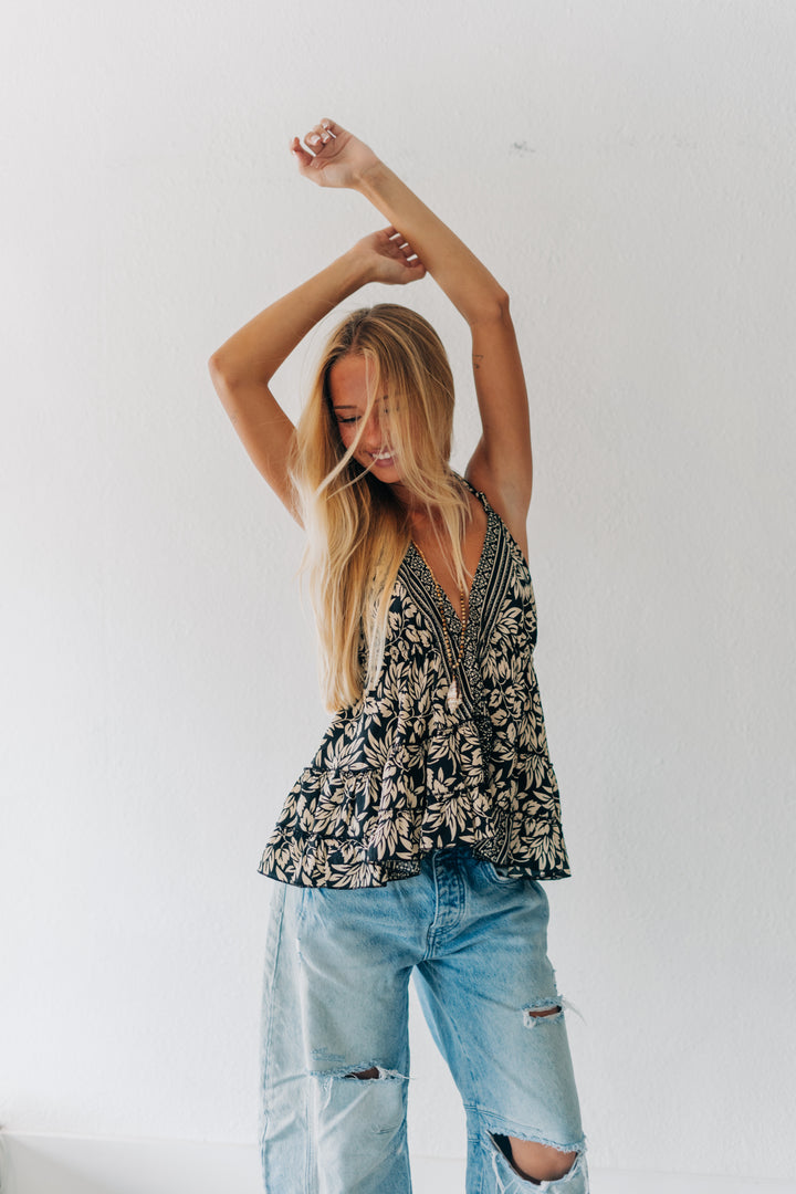 A woman with long blonde hair poses in a black and white botanical print sleeveless top and light-wash distressed jeans against a plain white wall.