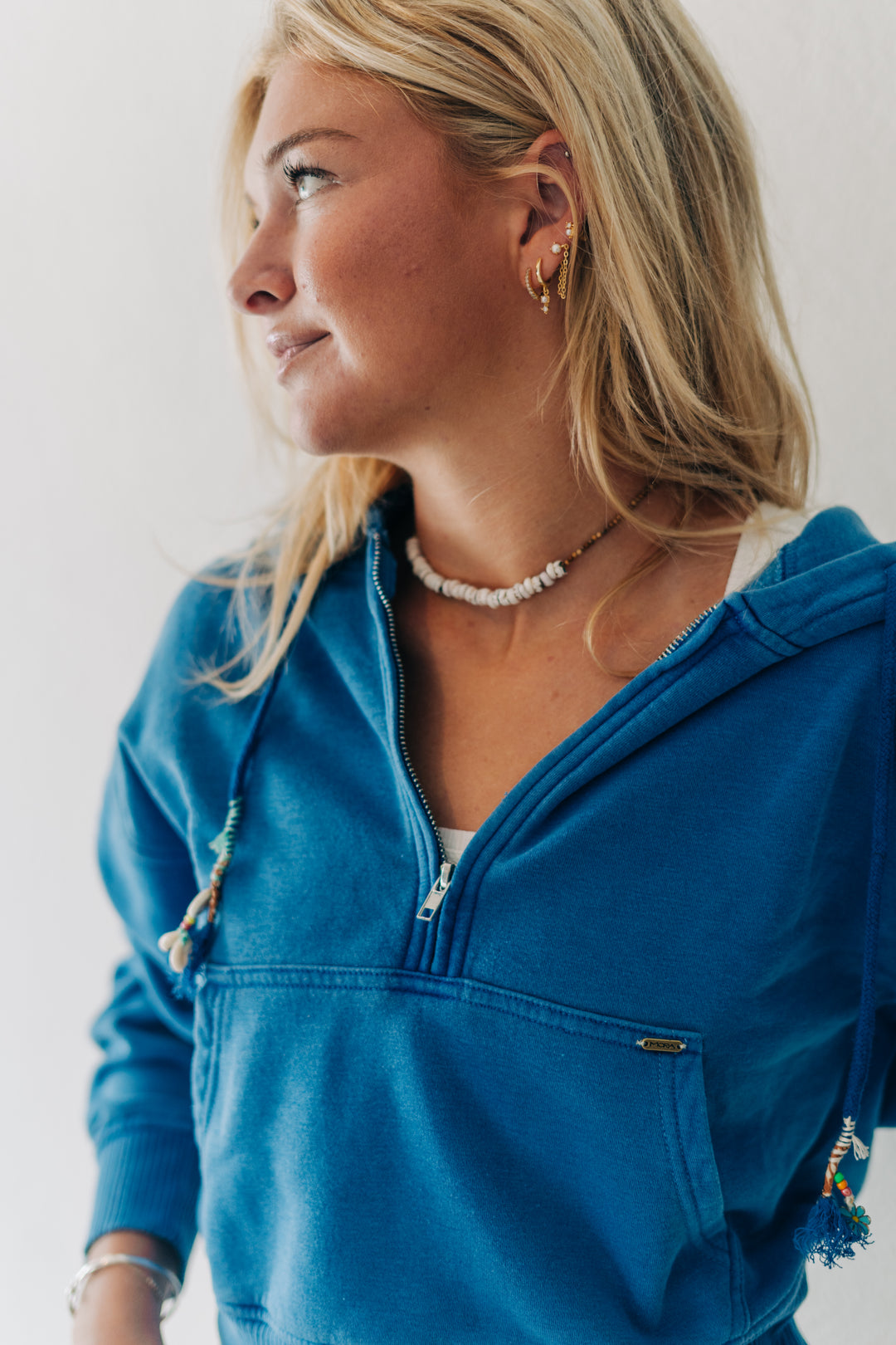 A woman wearing a bright blue zip-up hoodie with drawstrings and tassel details, shown in a close-up profile shot against a plain light background.