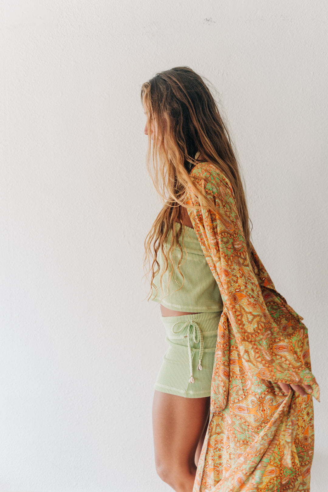 A woman stands in profile against a plain white wall, wearing a light sage green crop top and matching shorts, with a long flowing orange paisley-patterned shawl draped over her arm.