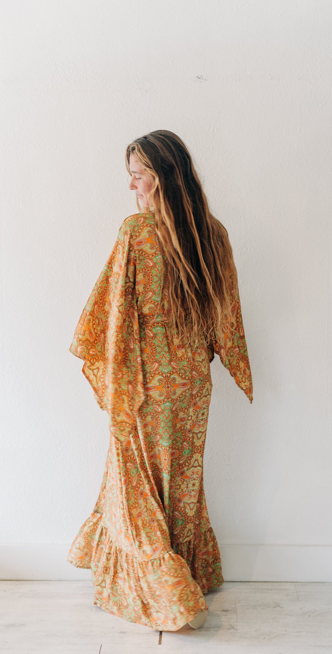 A woman with long light brown hair stands with her back to the camera, wearing a flowing, loose-fitting maxi dress with wide sleeves. The dress features an intricate paisley pattern in orange, yellow, and green.
