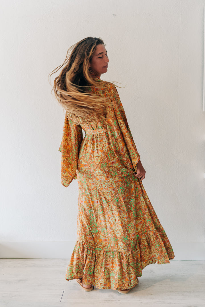 A woman wearing a long-sleeved, high-waisted maxi dress with a vibrant orange, yellow, and green paisley pattern, standing in profile against a plain white wall.