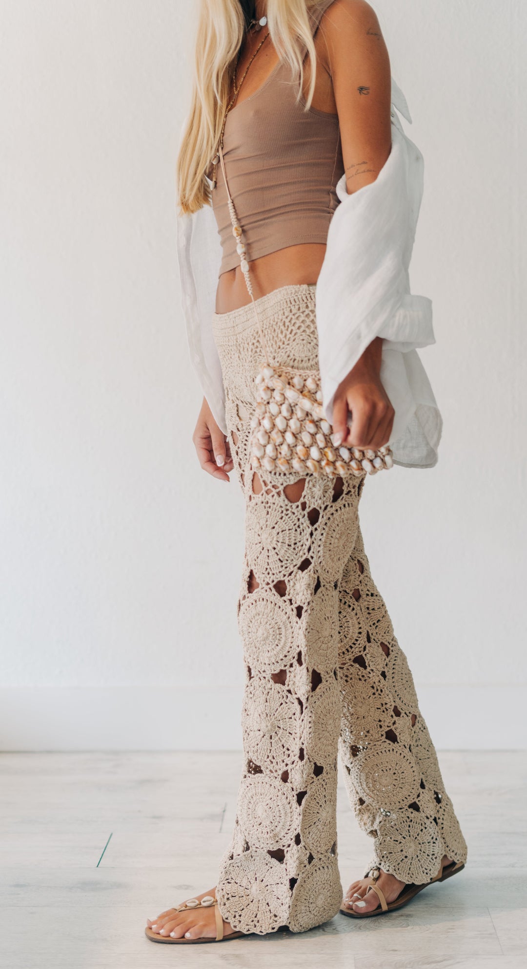 A woman modeling a pair of beige crochet flare pants with cutout details, paired with a brown crop top, white open-front cardigan, and a pearl-embellished clutch, set against a minimalist white wall and light floor.
