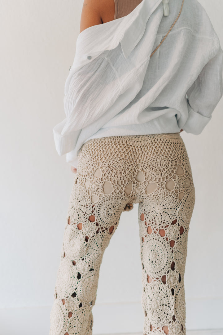 A person shown from the back wearing beige crochet lace pants and a light off-white draped shirt against a plain white wall.