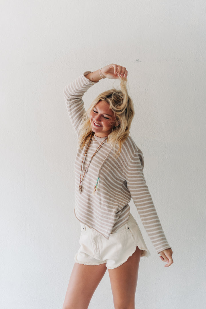 A woman posing in a beige and white striped long-sleeve crop top and white shorts against a plain white wall.
