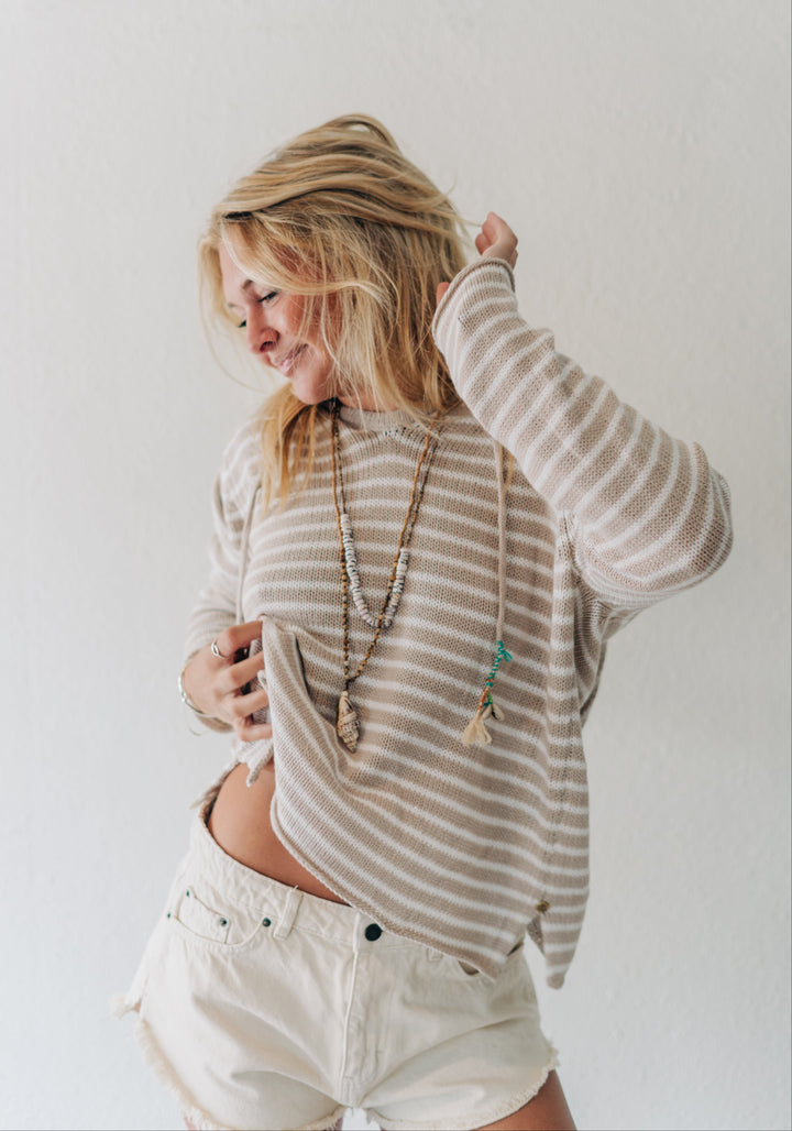 A woman with blonde hair wearing a beige and white striped long-sleeve knit top and cream shorts, standing against a plain white wall.