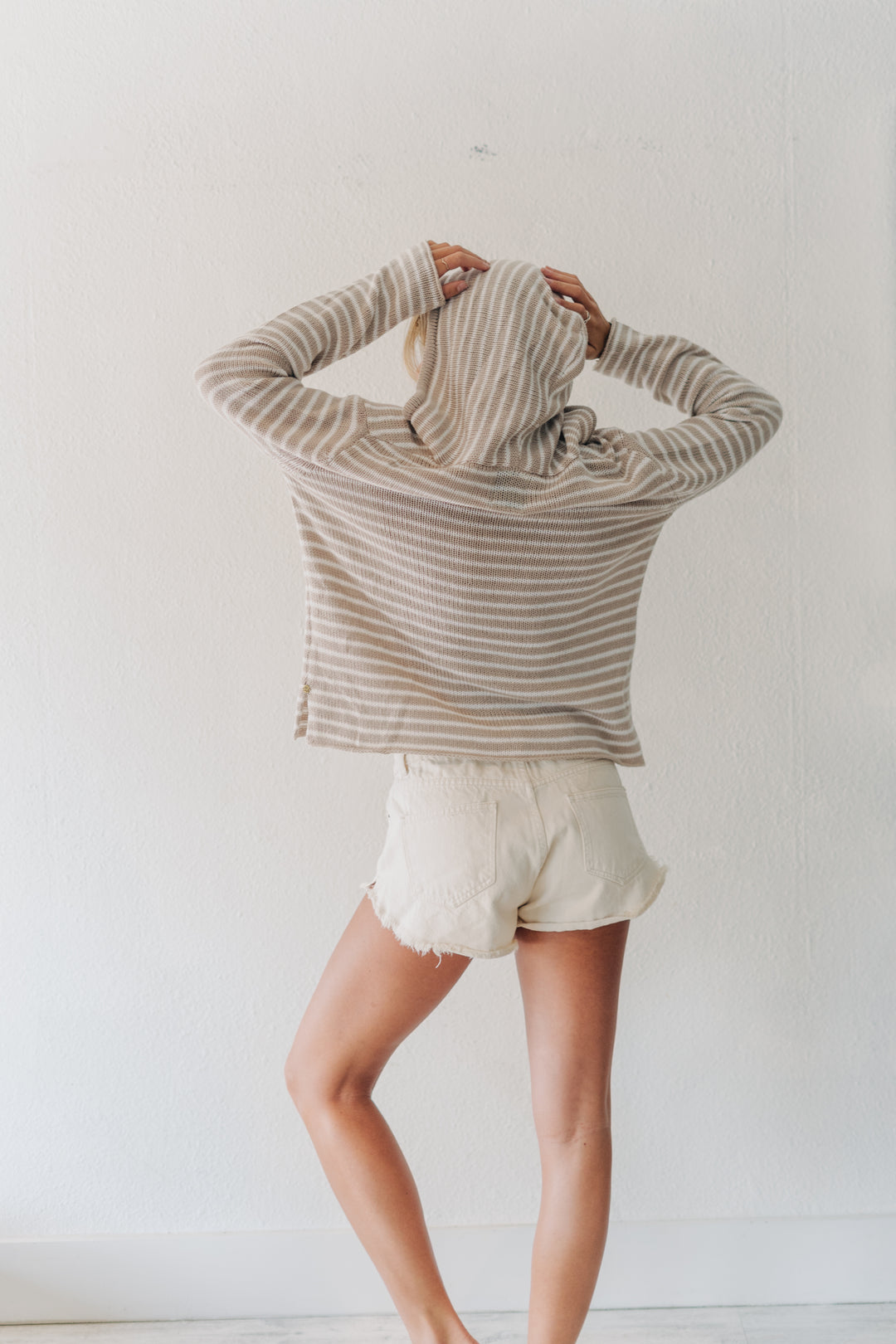 A person standing with their back to the camera, wearing a beige and white horizontally striped long-sleeve hooded sweatshirt and off-white shorts against a plain white wall.