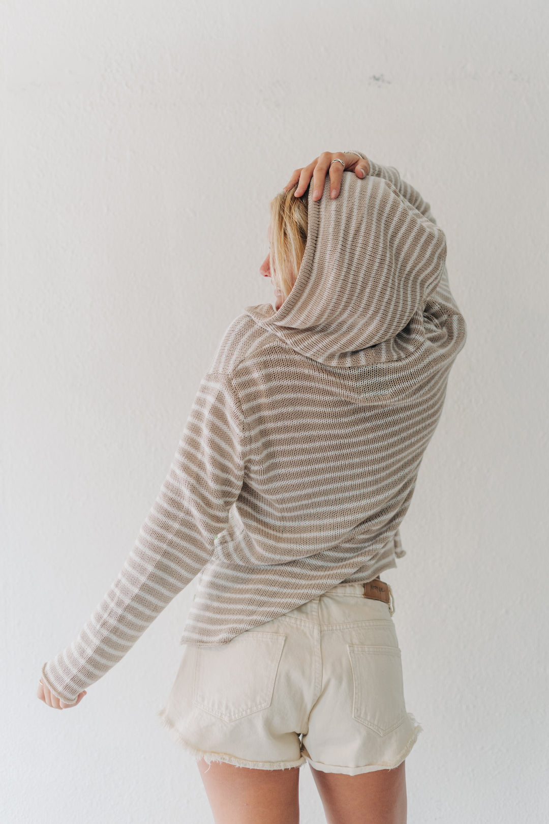 A woman shown from the back wearing a beige and white horizontally striped long-sleeve hoodie with a cowl neck, paired with cream-colored denim shorts, against a plain white wall.