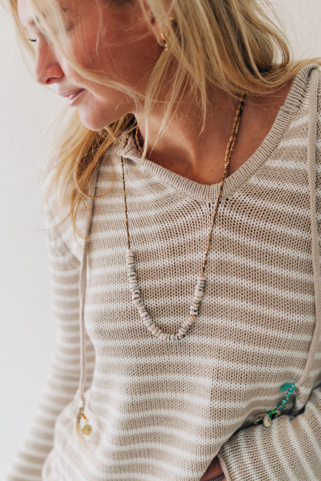A woman wearing a beige and white horizontally striped knit sweater, styled with layered necklaces and bracelets, set against a plain white wall.