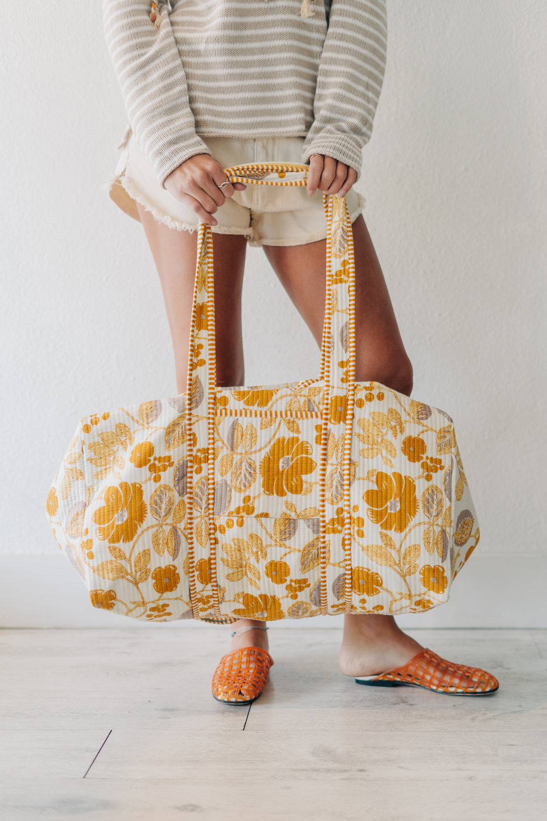 A person holding a large cream-colored duffel bag with a bold yellow floral pattern and matching yellow handles. The person is wearing a beige and white striped long-sleeve top, beige shorts, and orange-and-white checkered slip-on shoes, standing against a plain white wall on a light wood floor.