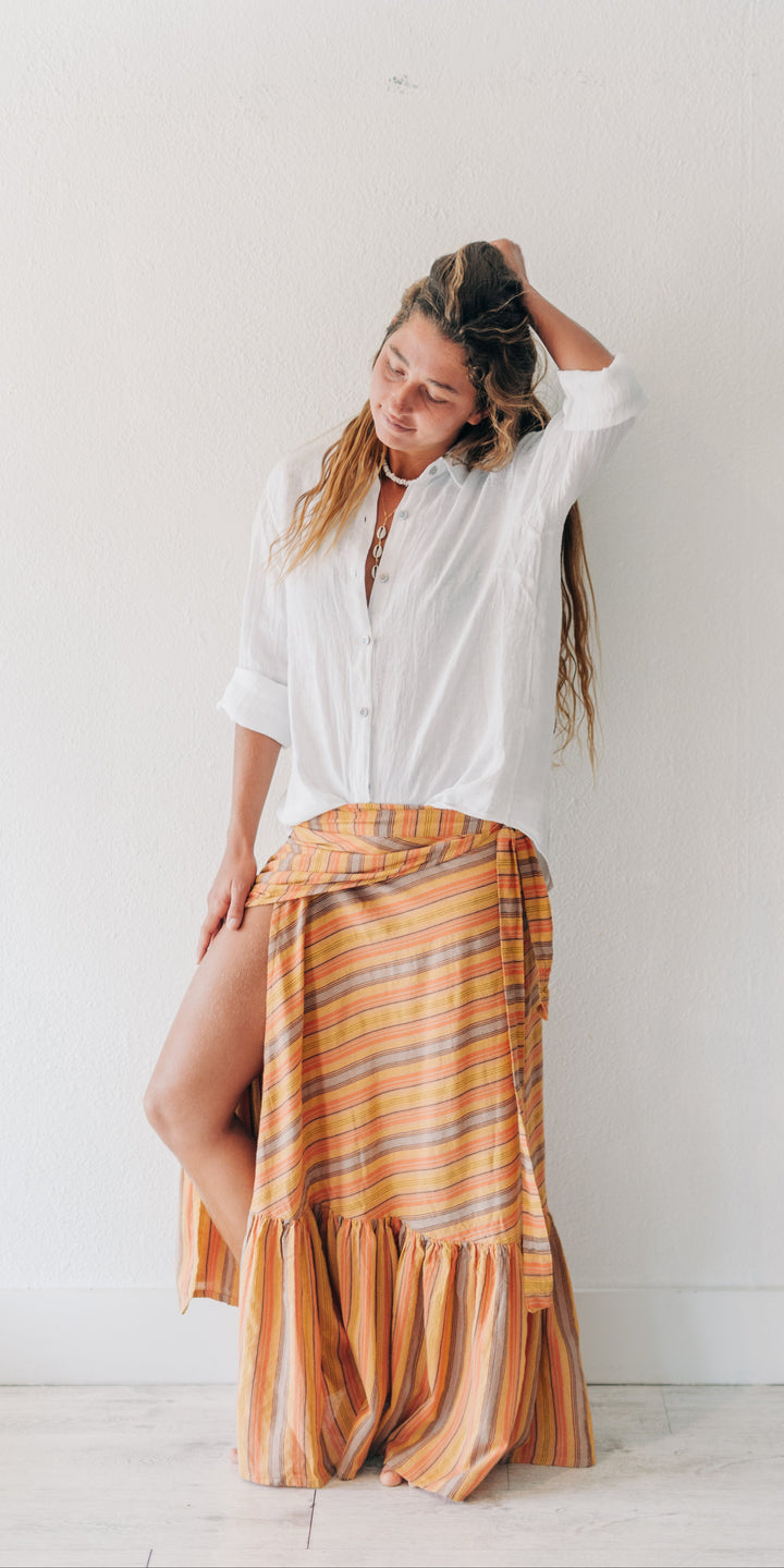 A woman posing in a white button-down shirt and a rust-orange striped wrap skirt with a ruffled hem, standing against a white wall.