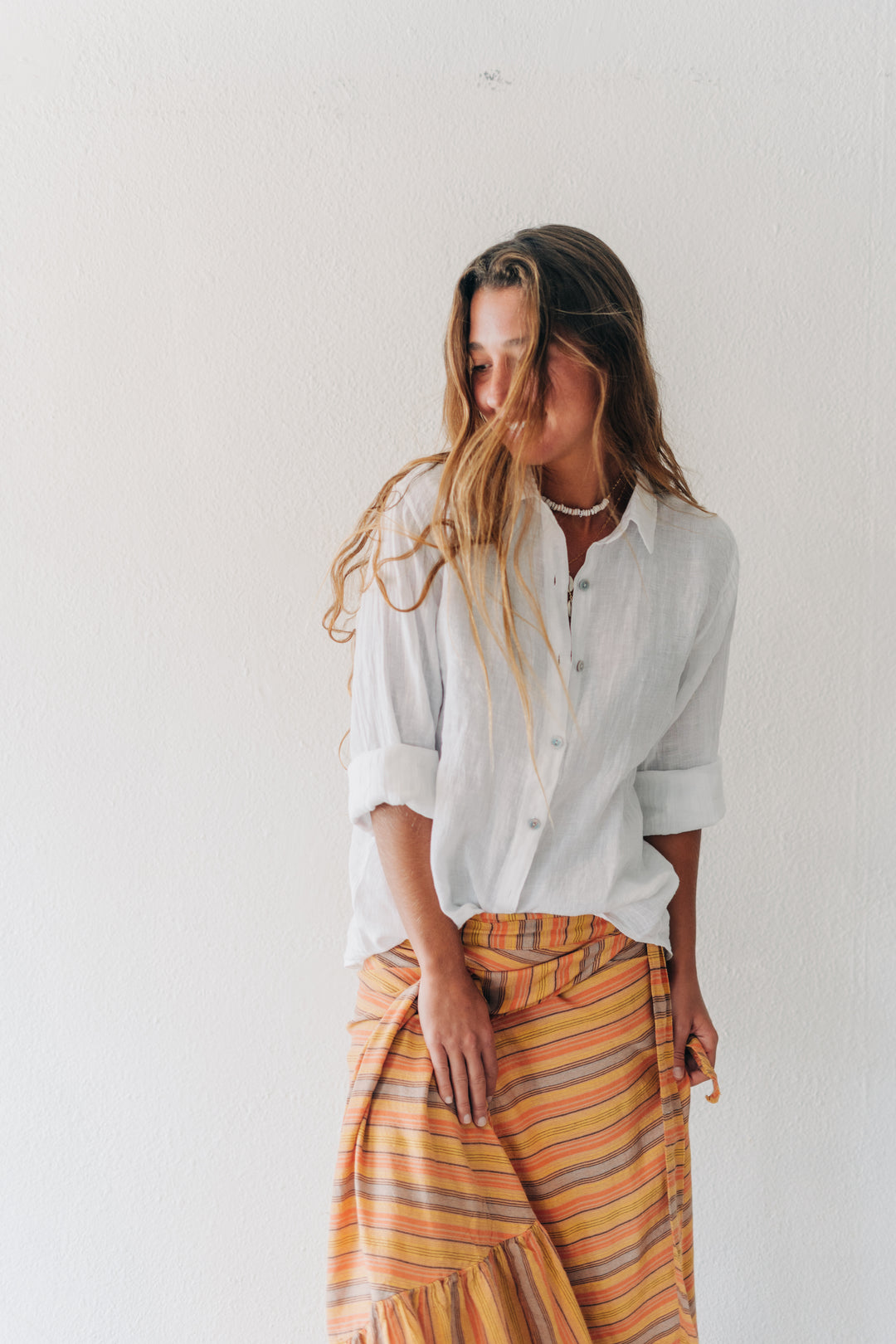 A woman wearing a white linen button-up shirt and a yellow, orange, and brown striped sarong skirt, standing against a white wall.