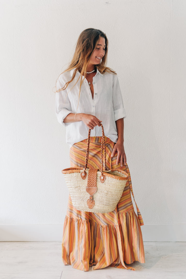 A woman stands against a plain white wall wearing a white button-up shirt and a long orange, yellow, and brown striped ruffled skirt, holding a large woven straw tote bag with brown leather handles.