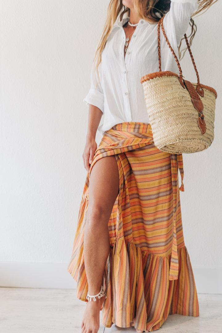A woman wearing a white button-up shirt and a high-low wrap maxi skirt with orange, yellow, and brown stripes. She is carrying a woven straw tote bag with brown leather handles against a plain white wall.