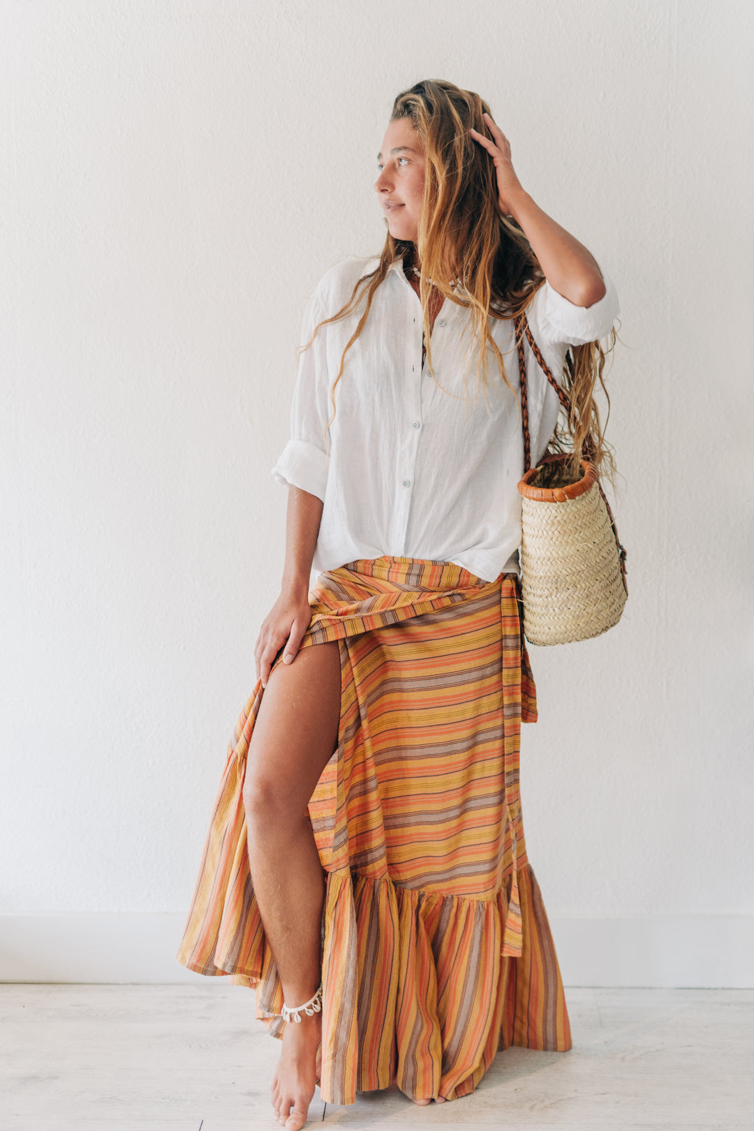A woman posing in a white button-down shirt and a vibrant orange, yellow, and brown striped ruffled wrap skirt, carrying a woven straw bucket bag against a plain white wall.