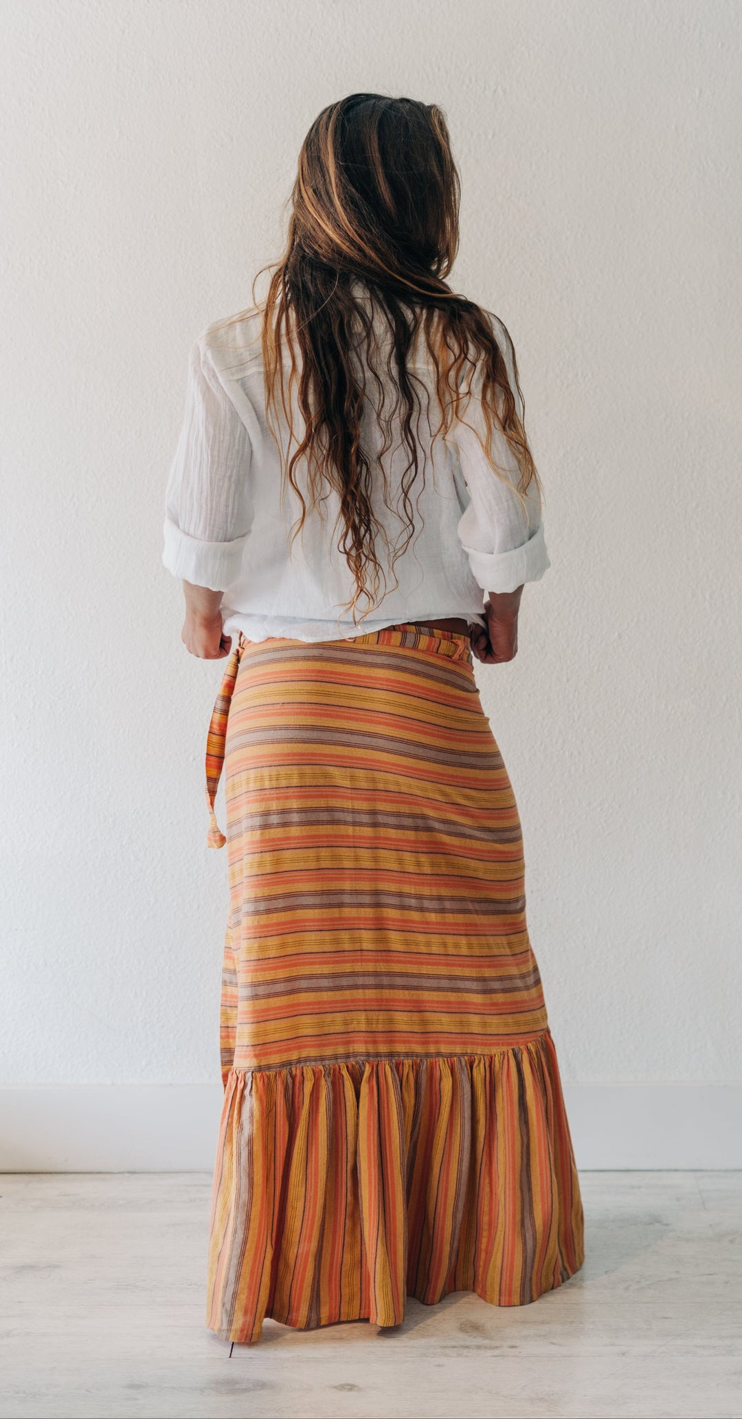 A woman viewed from behind wearing a white blouse and a long orange striped skirt with a ruffled hem, standing against a plain white wall.