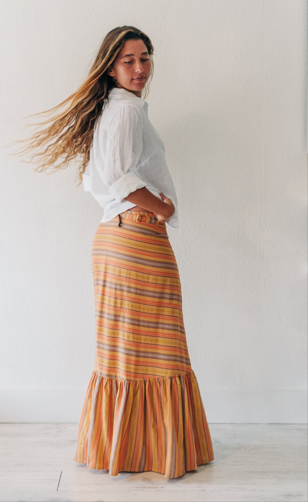 A woman wearing a white blouse and a long, ruffled striped skirt in orange, yellow, and brown tones, standing in a bright white room.