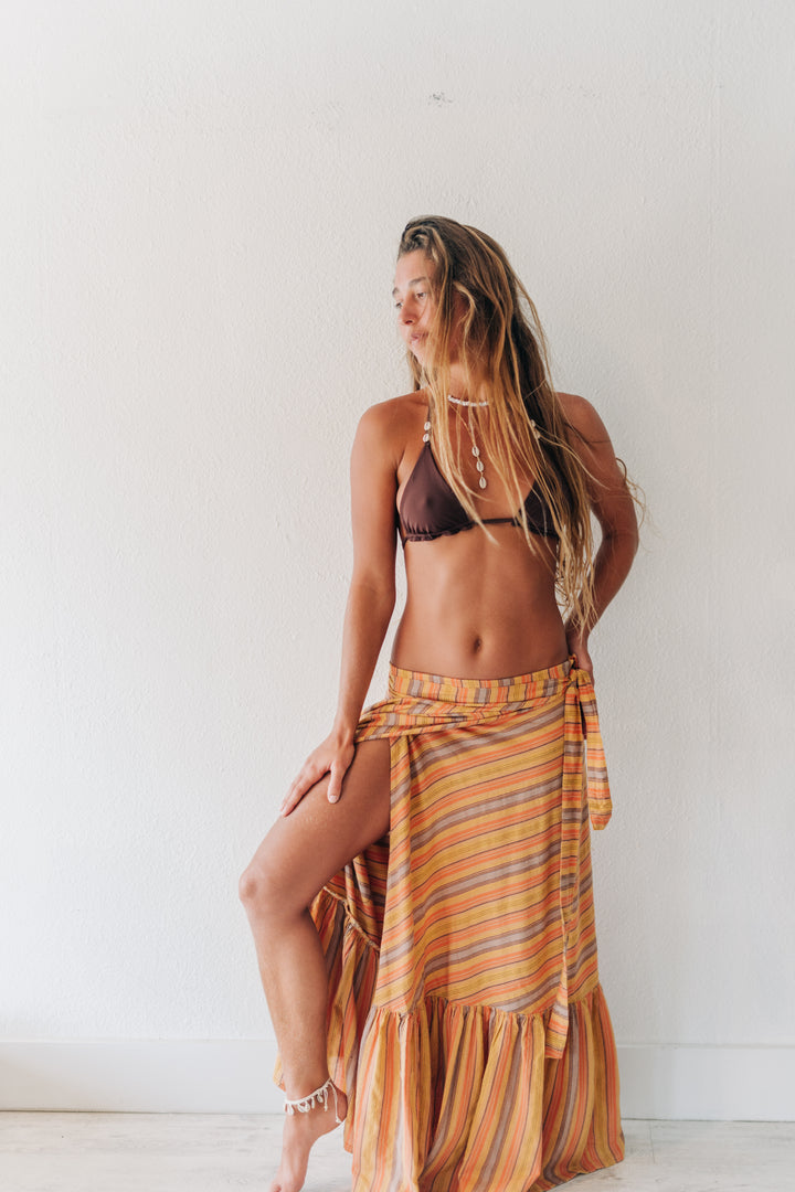 A woman posing in a striped orange and yellow sarong wrap skirt with a dark brown bikini top, standing against a plain white wall.