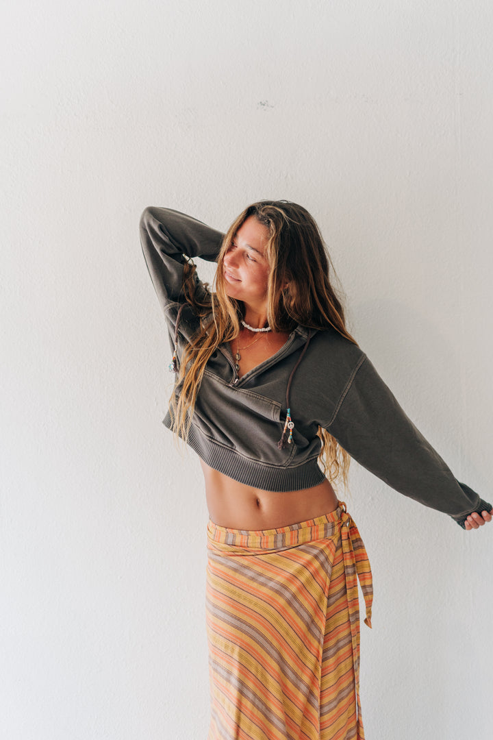 A woman posing against a white wall wearing a dark gray cropped zip-up hoodie and a striped orange, yellow, and brown sarong skirt.
