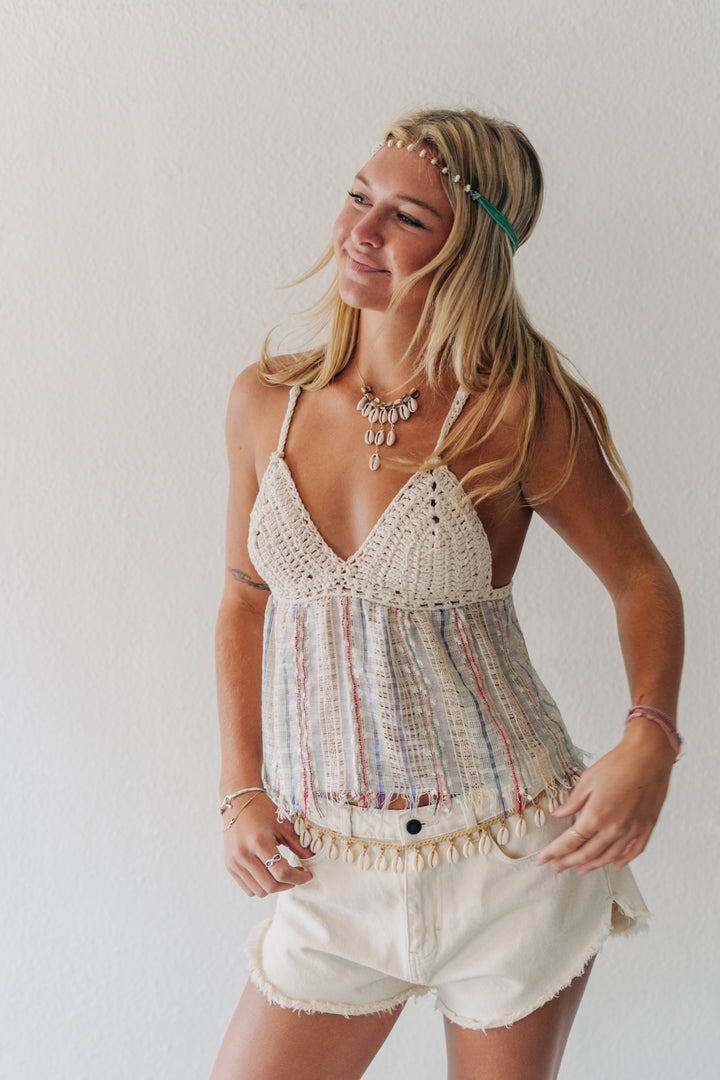 A woman modeling a bohemian-style crochet and striped camisole top with fringed trim, paired with cream shorts, against a plain white wall.
