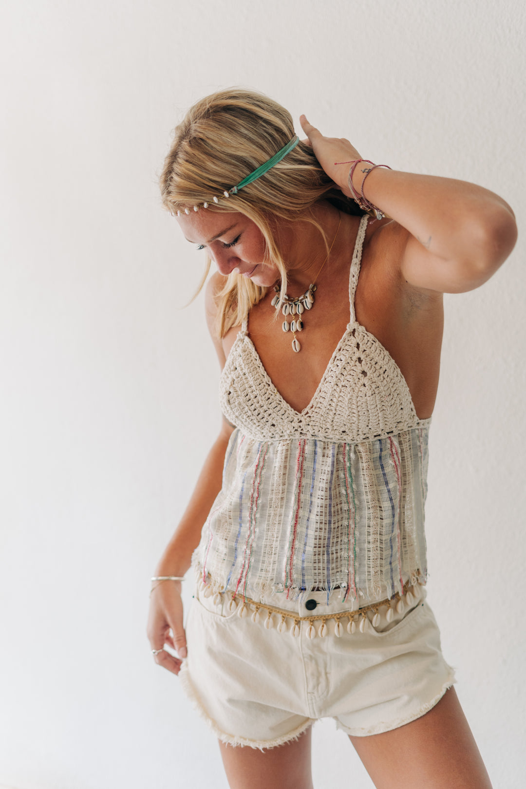 A woman wearing a cream crochet halter top with multicolored striped fringe and matching fringed shorts, styled with a beaded headband and statement jewelry against a plain white background.