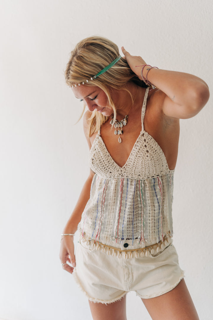 A woman wearing a cream crochet halter top with multicolored striped fringe and matching fringed shorts, styled with a beaded headband and statement jewelry against a plain white background.