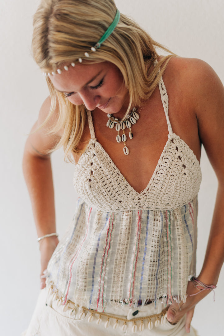 A woman wearing a cream crochet halter top with a fringed, striped woven overlay and white pants, accessorized with a shell necklace and a pearl headband.