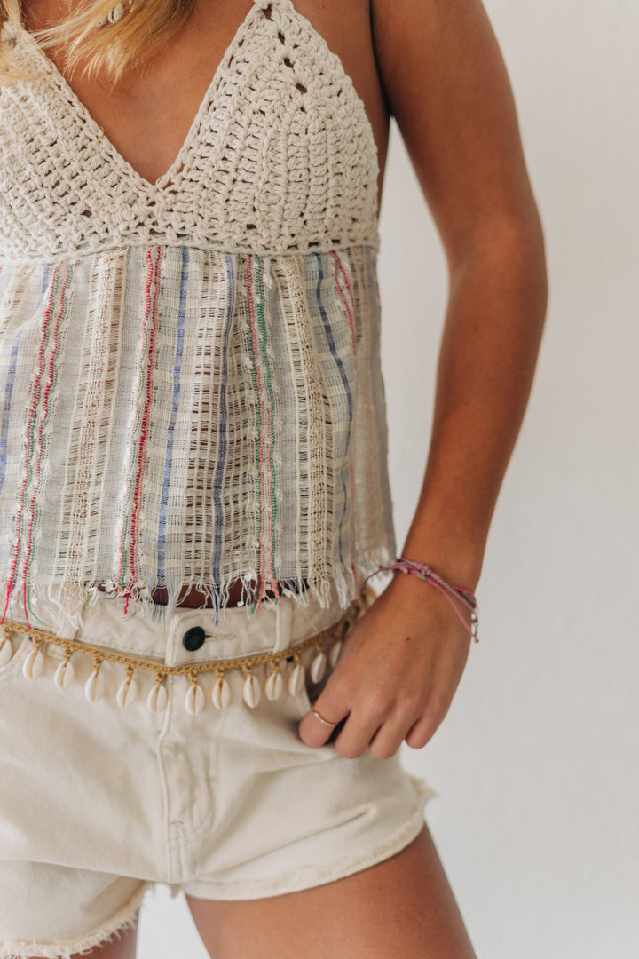 A close-up of a person wearing a crochet and woven striped tank top with fringe trim, paired with light denim shorts, against a plain light background.