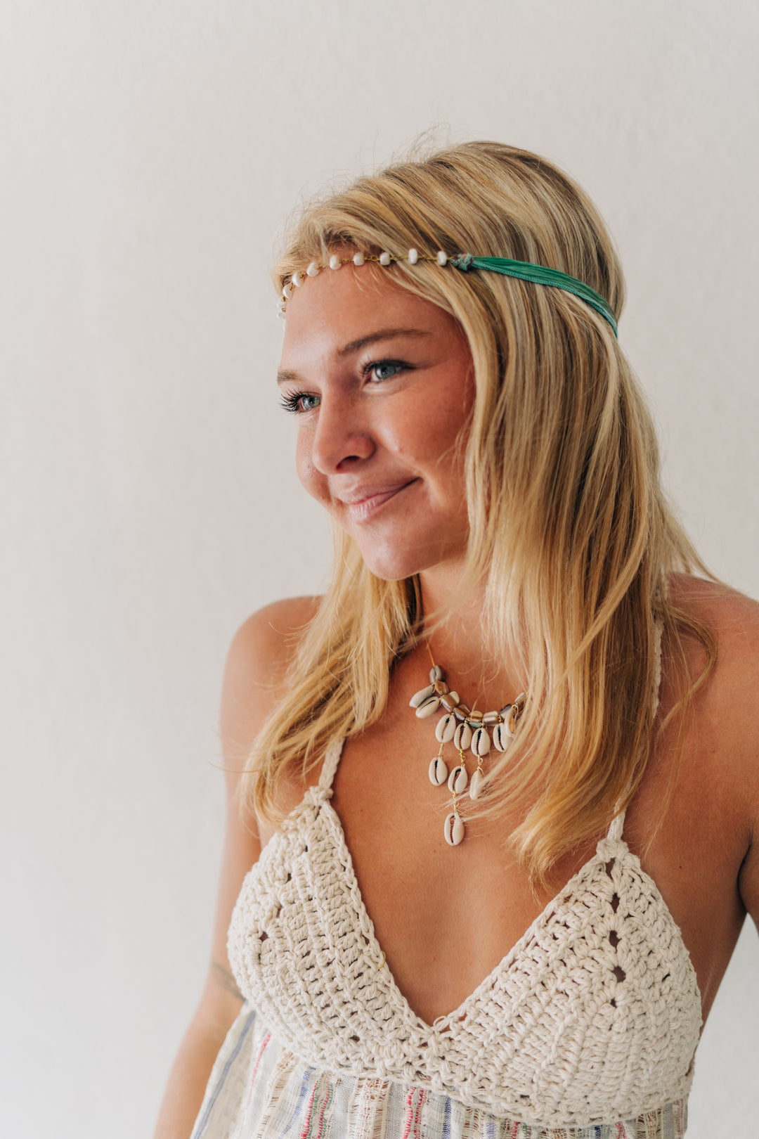 A woman with blonde hair wearing a green beaded headband and a bohemian-style crochet halter top, photographed against a plain off-white wall.