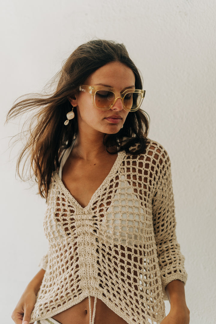 Woman wearing a beige crochet cover-up and sunglasses against a plain background
