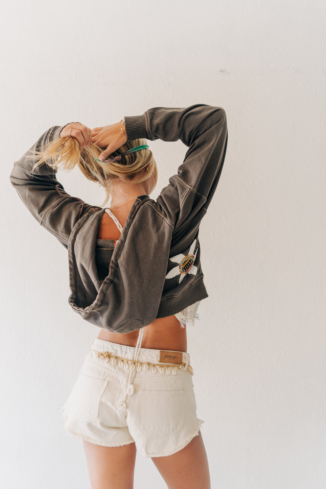 A woman with blonde hair in a ponytail, viewed from behind, wearing a dark brown cropped sweatshirt with a white graphic on the back and white denim shorts, posing against a plain white wall.