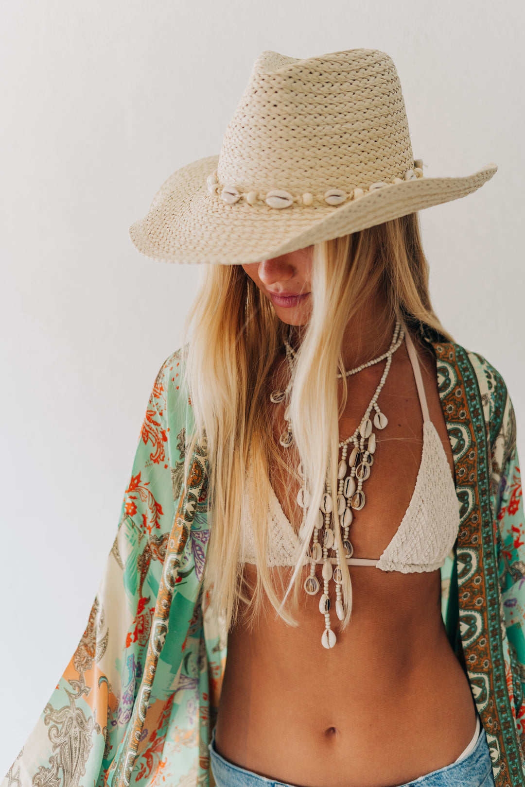 A woman wearing a beige woven cowboy hat with shell accents, a white crochet bikini top, layered silver shell necklaces, and a green and orange patterned open-front cover-up against a plain white background.