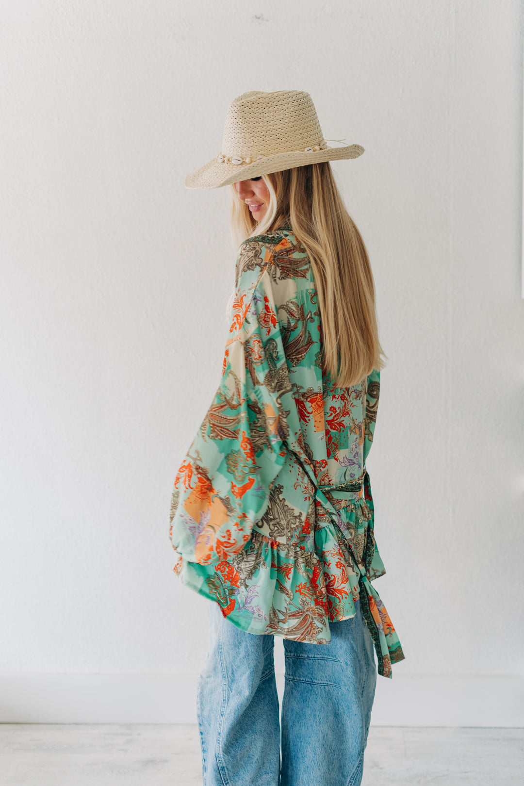 A woman wearing a light beige straw hat with gold accents, a green and orange tropical print wrap shirt, and blue jeans, standing against a white wall.