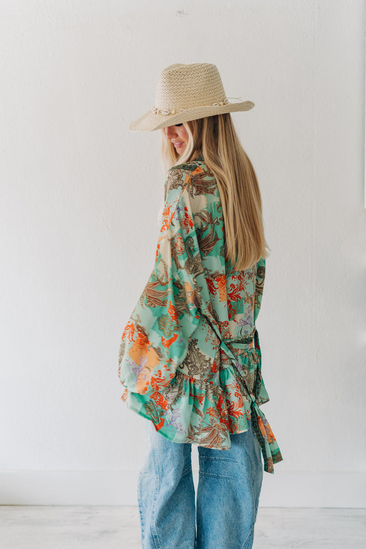 A woman wearing a light beige straw hat with gold accents, a green and orange tropical print wrap shirt, and blue jeans, standing against a white wall.