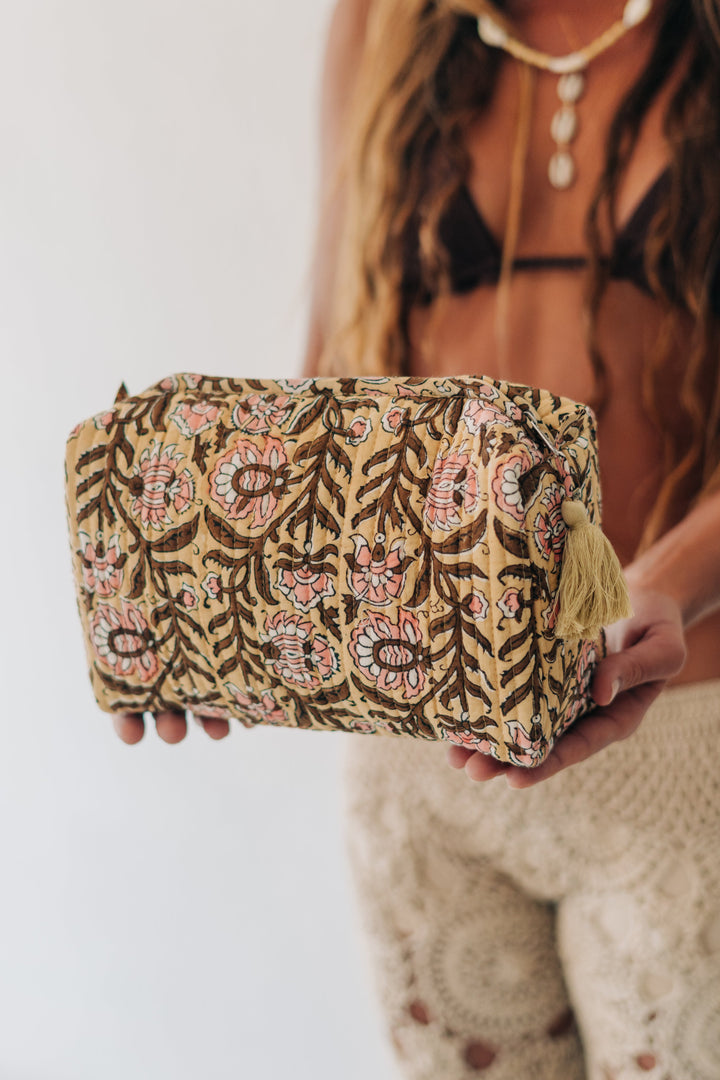 A woman holds a rectangular cosmetic pouch with a brown, beige, and pink floral patterned fabric cover and a small gold tassel zipper pull, against a bright white background.