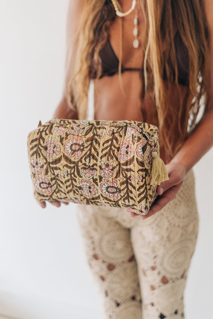 A woman holds a small rectangular pouch or clutch with an ornate bohemian-style floral and paisley pattern in beige, brown, pink, and cream. The pouch has a tassel detail on the side and is shown against a bright white background.