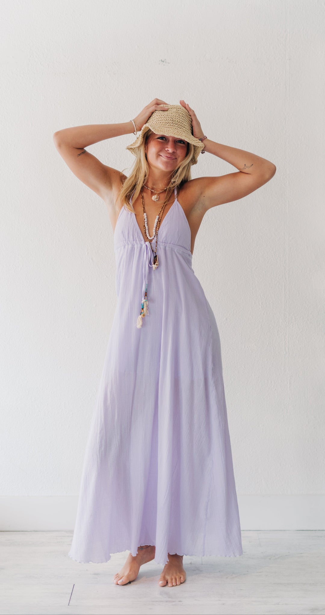 A woman wearing a light lavender halter maxi dress, a straw hat, and layered necklaces stands barefoot in a bright, minimalist white room.