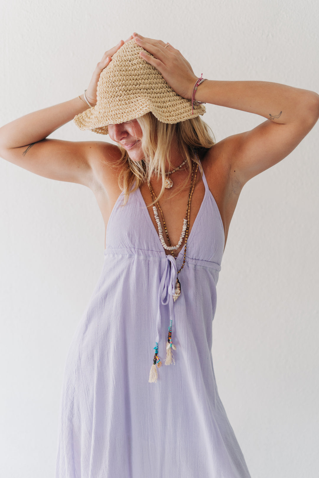 A woman wearing a light lavender halter dress and a straw sun hat, posing with her hands on her head against a plain white wall.