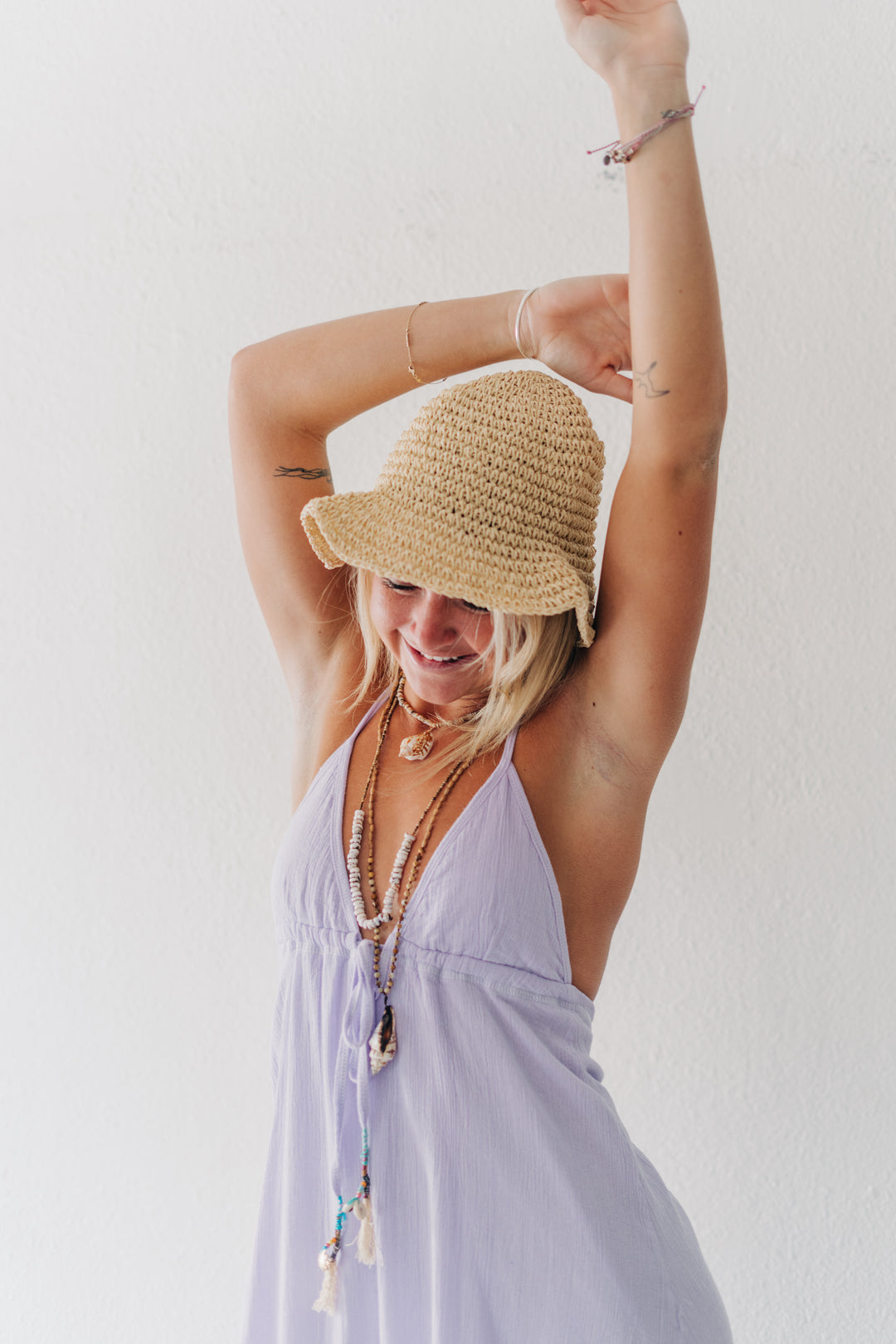 A woman wearing a light purple halter dress and a straw sun hat, posing with her arms raised against a plain white wall.