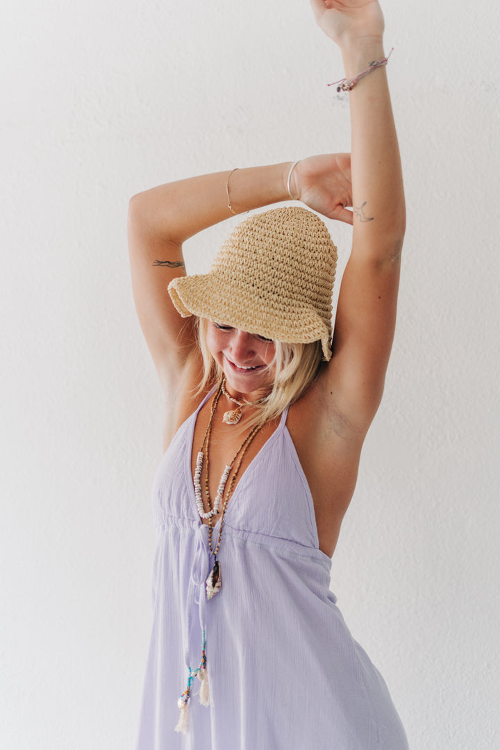 A woman wearing a light purple halter dress and a straw sun hat, posing with her arms raised against a plain white wall.