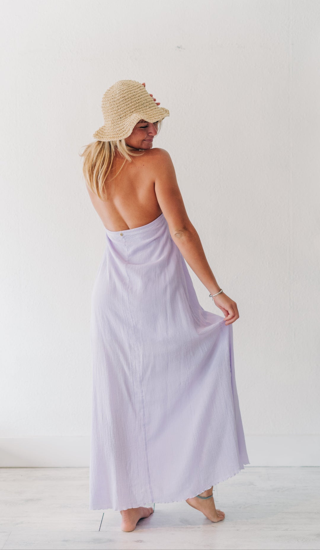 A woman wearing a light lavender maxi dress and a straw sun hat, shown from behind in a bright white room.