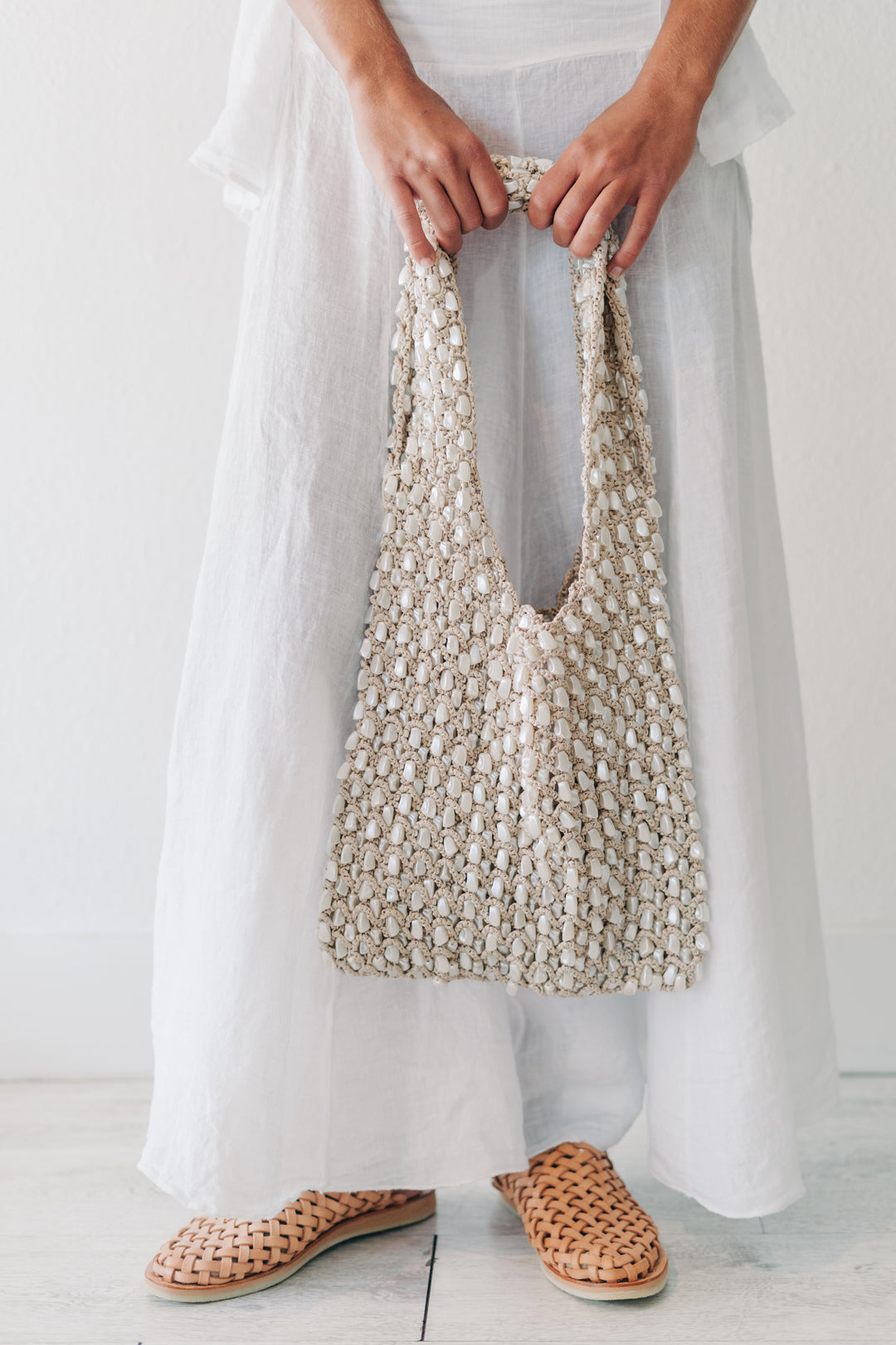 A person wearing a long white dress and woven tan slip-on shoes holds a large chunky knit shoulder bag in cream and beige.